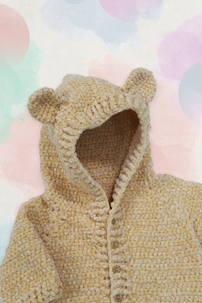 Círculo Amigurumi Pelúcia – Polyester Chenille Yarn (131 m / 85 g) - A close-up of a beige chunky knit hooded cardigan with bear ears on the hood and front buttons, shown against a pastel watercolor background.