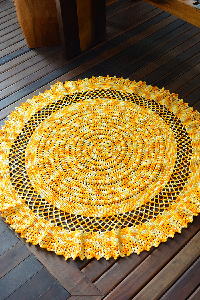 Circulo Barroco Multicolor Premium 4/6 - Brazilian Cotton Yarn, 226m/200g - A round yellow crocheted placemat with an intricate lace-like scalloped border, displayed on a dark brown wooden deck.