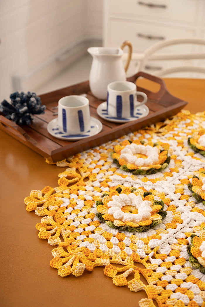 Circulo Barroco Multicolor Premium 4/6 - Brazilian Cotton Yarn, 226m/200g - A yellow and white crocheted table runner with floral motifs and a scalloped edge, styled on a wooden table next to a matching crocheted coaster with a white and blue ceramic cup and saucer set on a wooden tray.