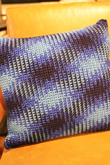 Circulo Barroco Multicolor Premium 4/6 - Brazilian Cotton Yarn, 226m/200g - A square crocheted throw pillow with a textured chevron pattern in shades of purple, blue, and dark brown, resting on a warm orange-brown leather sofa.