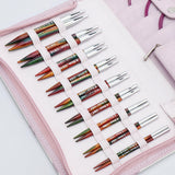 KnitPro Symfonie 2 Special interchangeable needle set, 10 cm, Deluxe - A pink fabric needle case holding multiple knitting needles arranged in elastic loops. The needles have silver metal tips and colored shafts in green, orange, red, and brown tones, with some white text printed on them.